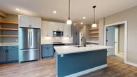 Located in Mount Pleasant, Wisconsin, this two-story home showcases a thoughtfully designed interior with modern finishes throughout. The kitchen serves as a standout feature, combining white upper cabinetry with slate-blue lower cabinets and a matching large center island equipped with a undermount sink and pendant lighting overhead. White subway tile backsplash, stainless steel appliances—including a French-door refrigerator, range, and built-in microwave—and open wood shelving complete the well-equipped space. Light wood-tone flooring extends from the kitchen into the open living area, which features neutral gray walls and a clean, versatile layout. Bedrooms are carpeted in a soft gray tone with large windows fitted with white blinds, allowing ample natural light. Bathrooms are finished with light tile flooring, gray vanities, white quartz countertops, and chrome fixtures; one bathroom includes a walk-in glass-door shower. An attached garage with concrete flooring and an automatic opener provides convenient parking and storage. The overall aesthetic is contemporary and cohesive, with a neutral palette accented by modern hardware throughout.