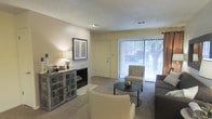 This single-floor residence offers a comfortable and inviting layout designed for relaxed living. The living area serves as the heart of the home, centered around a classic fireplace framed by warm, neutral-toned walls and soft carpeted flooring that adds a sense of coziness throughout. A decorative cabinet with ornate detailing sits beside the fireplace, providing both style and storage, while framed wall art contributes to the room's welcoming ambiance. Natural light floods the space through a large sliding glass door dressed with vertical blinds, offering a view of mature trees just outside and creating a seamless connection between the interior and the outdoors. The living area is thoughtfully arranged with a sofa, accent chairs, and a round coffee table, making it an ideal space for both relaxing and entertaining. Recessed lighting in the textured ceiling provides additional illumination, complementing the warm glow of table lamps placed throughout the room. The overall atmosphere is calm and comfortable, with a neutral color palette and tasteful furnishings that enhance the sense of space and openness throughout this single-level home.
