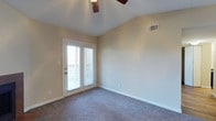 This single-floor space features a well-appointed room highlighted by vaulted ceilings that lend an open, airy feel to the interior. The ceiling is fitted with a ceiling fan, providing both practical air circulation and a subtle decorative element. Neutral beige walls create a versatile backdrop, complementing the plush wall-to-wall carpeting that adds warmth and comfort underfoot. A standout feature of the room is the set of French doors, framed in white and fitted with slatted blinds, which flood the space with natural light while providing direct access to an outdoor area. The doors offer a seamless connection between the interior and exterior, enhancing the sense of openness. An HVAC vent is positioned on the upper wall, ensuring efficient climate control throughout. White baseboards provide clean, crisp trim detail against the neutral walls. The overall ambiance is bright, calm, and move-in ready, with a straightforward layout that allows for flexible furniture arrangements and easy personalization.