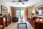 This single-floor residence offers a thoughtfully designed layout with distinct living, dining, and sleeping areas. The living room serves as the heart of the home, featuring plush wall-to-wall carpeting, a ceiling fan with light fixture, and a sliding glass door that opens to a private balcony overlooking lush greenery, allowing natural light to fill the space. A dedicated dining area sits adjacent to the living room, complete with a pendant light fixture and ample room for a full dining set. The galley-style kitchen is efficiently arranged with warm wood cabinetry, light countertops, a double sink, and hardwood-style flooring, providing functional storage and prep space. The primary bedroom is a comfortable retreat with carpet flooring, a ceiling fan, and room for a full furniture arrangement. A second bedroom offers closet storage and convenient proximity to an in-unit laundry area. The bathroom is finished with full tile surrounds and a soaking tub-shower combination. Neutral tones and warm finishes are carried throughout the space, creating a cohesive and inviting ambiance from room to room.