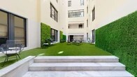 This contemporary building centers around a striking open-air courtyard that serves as its social and architectural focal point. Enclosed by multi-story cream-colored facades with large black-framed windows and balconies, the courtyard is flooded with natural light from above, creating an open and refreshing atmosphere. The ground level is finished with lush artificial turf, providing a clean and vibrant surface that contrasts beautifully with the surrounding architecture. Vertical green walls line multiple sides of the courtyard, adding a bold, natural element to the modern design. Scattered throughout the space are wire-frame chairs and café-style tables, offering a relaxed setting for casual gatherings or informal meetings. Marble-finished steps lead into the courtyard from at least one end, lending an elegant touch. An upper-level balcony, framed by sleek glass and brass railings, overlooks the courtyard below and extends the usable social space vertically. Interior corridors feature polished terrazzo flooring and recessed ceiling lighting, maintaining a clean, professional aesthetic throughout. Together, these elements create a well-balanced environment that blends biophilic design with modern architecture, making the courtyard a welcoming hub within the building.