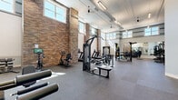 Located at 500 N Henry in Williamsburg, Virginia, this single-floor fitness center offers an expansive and well-appointed exercise facility across two distinct exercise rooms. The main workout area is an impressively large, open space characterized by soaring ceilings, exposed brick columns, and generous natural light streaming through rows of elevated windows. Durable rubber flooring covers the entire area, and the room is outfitted with a comprehensive selection of strength and cardio equipment, including cable machines, weight racks, kettlebells, medicine balls, treadmills, and stationary bikes. Full-length mirrors line one wall, enhancing the sense of space. Decorative trim detailing along the upper walls adds architectural character, while a motivational neon sign and a wood-slat accent wall serve as focal design elements. A staircase with modern metal railings connects to an upper-level overlook, adding vertical drama to the space. The second exercise room offers a quieter, studio-style environment finished in cool muted tones. It features a smooth light-colored floor, ceiling fans, wall-mounted mirrors, a flat-screen television, and equipment suited for cycling, stretching, and functional training. Together, the two rooms create a comprehensive, well-designed fitness facility.