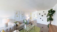 This single-floor space offers a clean, contemporary aesthetic defined by bright white walls, smooth polished concrete floors, and an open, airy layout. The living area serves as the centerpiece of the space, furnished with a tufted light gray sofa accented by sage green and neutral throw pillows. A glass-top coffee table with a gold-toned frame sits atop a green area rug, adding a pop of color and texture to the room. Two large floral canvas prints hang above the sofa, contributing an elegant, gallery-like touch to the wall. Decorative elements, including a sculptural wall installation and a tall white vase with greenery, enhance the space's curated, modern feel. A sleek side table with a white table lamp provides warm ambient lighting. The open layout flows seamlessly toward what appears to be a dining area and kitchen in the background, where natural light filters through a window, brightening the entire space. The overall ambiance is light, fresh, and inviting, combining modern furnishings with soft organic accents to create a balanced and welcoming interior environment.
