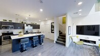 This two-story space showcases a thoughtfully designed open-concept layout that seamlessly connects the kitchen, dining, and living areas across its main level. The kitchen is a standout feature, anchored by a striking navy blue island with a white countertop and complemented by dark cabinetry, stainless steel appliances including a French door refrigerator, and pendant lighting overhead. Small potted plants along the upper cabinets add a touch of greenery to the space.

Light grey laminate flooring runs throughout the main level, providing a cohesive and contemporary foundation. The living area benefits from impressive double-height ceilings with a skylight or upper window that draws natural light deep into the space, creating an open and airy atmosphere. Bold, graphic wall art in green and white tones adds visual interest and ties together the color palette seen throughout the home.

A carpeted staircase with clean white walls leads to the upper floor, while framed artwork and accent chairs with chartreuse upholstery contribute to the modern, curated aesthetic. Recessed lighting throughout ensures the space remains well-lit and inviting at all hours.