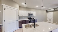 This single-story unit offers a thoughtfully designed layout across a compact but efficient footprint. The open-concept kitchen and living area serves as the heart of the space, anchored by a central island with a double stainless steel sink, dark espresso lower cabinetry, and crisp white upper cabinets. Light-toned countertops and a matching backsplash provide a clean contrast. Stainless steel appliances, including a range, microwave, and refrigerator, complete the kitchen. Glass pendant lights hang above the island, while recessed lighting illuminates the space throughout. Light luxury vinyl plank flooring runs through the main living and kitchen area, adding a modern, low-maintenance finish.

Both bedrooms feature plush carpeting and neutral gray walls, with exposed industrial-style ductwork running along the ceilings — a distinctive architectural detail. The primary bedroom includes a ceiling fan and a window providing natural light. The second bedroom connects directly to the main living area via a short hallway and includes a closet. The bathroom is clean and functional, outfitted with a walk-in shower, matte black hardware accents, and light tile flooring, maintaining a consistent contemporary aesthetic throughout the unit.