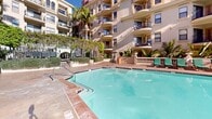 Located in the heart of downtown Los Angeles, this resort-style outdoor amenity space at 725 S Bixel Street offers a beautifully designed courtyard that blends Mediterranean-inspired architecture with sophisticated urban living. The centerpiece is a generously sized swimming pool with crystal-clear water, complemented by a separate spa and hot tub, both finished with elegant tilework and surrounded by stamped concrete and natural stone decking.

Framed by the multi-story building's ornate façade — featuring wrought-iron balconies, arched windows, decorative cornices, and classical balustrades — the courtyard exudes a refined, European ambiance. Rows of cushioned chaise lounges in vivid teal are arranged throughout the deck, offering ample space for sunbathing and relaxation beneath the abundant Southern California sunshine.

Lush tropical landscaping enhances the setting, with towering palm trees, banana plants, Italian cypress trees, and potted tropical foliage creating a verdant, secluded atmosphere. A pergola draped in climbing greenery provides shaded seating areas near the spa. The open-air terrace also offers dramatic views of the downtown Los Angeles skyline, including prominent high-rise towers visible above the treetops, seamlessly connecting this tranquil retreat to the vibrant city beyond.