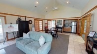 This single-story space is defined by its striking vaulted ceiling, which rises dramatically to create an airy, expansive atmosphere throughout the open-plan interior. A ceiling fan is centrally mounted at the apex, ensuring comfortable air circulation across the entire space.

The living area anchors one end of the layout, furnished with a comfortable light blue loveseat accented with decorative pillows and placed atop a large area rug over tile flooring. A dark wood armoire and a wall clock add character to this corner, alongside a water cooler that contributes to the space's functional appeal.

The dining area sits centrally, featuring a dark wood table with matching chairs, positioned to take advantage of natural light streaming through a pair of French doors. A second set of French doors nearby further enhances the bright, welcoming atmosphere and provides additional outdoor connectivity.

The kitchen occupies the far end of the space, offering warm wood cabinetry, a stainless steel refrigerator, and counter space with a coffee station. The consistent light tile flooring ties the entire open-plan layout together, making the space feel cohesive and well-organized throughout.