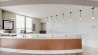 Located at 100 Myrtle St in Longwood, Florida, this single-floor community clubhouse offers a thoughtfully designed amenity space that blends contemporary style with functional comfort. The grand entrance lobby greets visitors with large-format tile flooring, a curved reception desk finished in warm wood paneling with a stone countertop, and elegant pendant lighting that sets a welcoming tone. Expansive floor-to-ceiling windows flood the interior with natural light and provide open views of the lush, landscaped surroundings. A generously sized open lounge area features carefully curated seating arrangements, vibrant abstract artwork adorning the walls, and recessed ceiling details that add architectural interest. A dedicated coffee and bar area serves as a social hub, complete with a mosaic-tiled bar front, stone countertops, pendant lighting, and built-in appliances. Adjacent to it, a bright dining area with round tables and teal upholstered chairs overlooks tropical palms and colorful plantings through oversized windows. Throughout the space, a cohesive neutral color palette is accented with teal, yellow, and warm wood tones, creating a vibrant yet sophisticated atmosphere ideal for community gatherings and everyday relaxation.