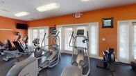 This fitness center is a well-appointed exercise facility designed to support a full range of workout routines. The space is immediately recognizable by its bold orange walls, which contrast sharply with the white trim, ceilings, and window frames to create an energetic and motivating atmosphere. Dark rubber flooring runs throughout, providing a durable, slip-resistant surface suitable for all types of exercise.

The cardio section is well-stocked, featuring multiple treadmills from brands including Spirit and True, elliptical trainers, and a recumbent stationary bike. These machines are positioned near large windows dressed with white shutters, allowing natural light to filter into the space. Wall-mounted flat-screen televisions are positioned throughout, offering entertainment during workouts.

A full-length mirrored wall spans one side of the room, visually expanding the space while providing users a clear view during strength training. A dedicated free weights area includes a well-organized dumbbell rack with a broad range of weights, alongside medicine balls and a weight bench. Additional strength and cable machines are situated toward the rear of the room. Overhead fluorescent lighting ensures the entire space is evenly and thoroughly illuminated throughout the day.