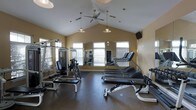This single-story fitness center offers a thoughtfully designed workout space beneath striking vaulted ceilings that lend an open, airy atmosphere to the room. Warm beige walls and multiple windows fitted with blinds allow abundant natural light to fill the space, creating a bright and inviting environment. Ceiling fans with pendant lights are suspended from the pitched ceiling, ensuring proper air circulation and comfortable lighting throughout.

The facility is outfitted with a comprehensive selection of exercise equipment, including multiple treadmills arranged along the back wall, a cable weight machine, a multi-functional strength training station, and various benches suitable for free weight exercises. A large wall mirror spans a significant portion of one side, enhancing the sense of space while allowing users to monitor their form during workouts. The flooring features durable, dark-toned material appropriate for a high-traffic fitness environment. A wall-mounted television is positioned for convenient viewing during cardio sessions. The overall layout is well-organized, providing ample room to move between stations comfortably.
