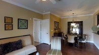 This single-floor residence exudes a warm and classic charm, thoughtfully designed with attention to detail throughout. The interior is unified by rich, dark hardwood flooring that flows seamlessly across the living and dining areas, grounding the space with a sense of warmth and sophistication.

The living area features a ceiling fan with overhead lighting and is adorned with elegant crown molding that frames the warm golden-yellow walls, lending a traditional and polished aesthetic. Tasteful artwork and decorative accents complement the neutral, upholstered seating, creating a comfortable and inviting atmosphere.

Adjacent to the living space, the dining area is anchored by a striking chandelier that casts a warm glow over an ornate dining table surrounded by upholstered chairs. A large decorative mirror enhances the sense of depth in the room, while a window allows natural light to filter in.

A wooden console table with candle accents and framed art serves as a graceful transition between the living and dining areas. The space also includes interior doors with classic paneling, one of which features louvered shutters, suggesting a storage closet. Overall, this home offers a refined, cohesive design that balances elegance with everyday comfort.