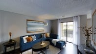 This single-floor residence offers a welcoming and well-appointed living environment with a clean, modern aesthetic. The living area serves as the heart of the space, showcasing a neutral gray color palette accented by warm tones that create an inviting atmosphere. A large sectional sofa anchors the room, complemented by a sleek oval coffee table and a plush area rug that adds texture and warmth to the hard flooring beneath. Coastal-themed artwork adorns the walls, reinforcing a relaxed, serene ambiance throughout. A textured ceiling adds architectural character overhead, while a stylish arc floor lamp provides ambient lighting. The room is bathed in natural light, thanks to expansive sliding glass doors that open directly onto a private balcony, offering outdoor views and a seamless connection between interior and exterior living. Sheer white curtains frame the glass doors, softening the natural light that filters in. The balcony features a classic railing and overlooks a landscaped outdoor setting. Overall, the space strikes a balance between comfort and contemporary style, making it a functional and visually appealing place to live.