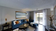 This single-floor residence offers a welcoming and well-appointed living environment with a clean, modern aesthetic. The living area serves as the heart of the space, showcasing a neutral gray color palette accented by warm tones that create an inviting atmosphere. A large sectional sofa anchors the room, complemented by a sleek oval coffee table and a plush area rug that adds texture and warmth to the hard flooring beneath. Coastal-themed artwork adorns the walls, reinforcing a relaxed, serene ambiance throughout. A textured ceiling adds architectural character overhead, while a stylish arc floor lamp provides ambient lighting. The room is bathed in natural light, thanks to expansive sliding glass doors that open directly onto a private balcony, offering outdoor views and a seamless connection between interior and exterior living. Sheer white curtains frame the glass doors, softening the natural light that filters in. The balcony features a classic railing and overlooks a landscaped outdoor setting. Overall, the space strikes a balance between comfort and contemporary style, making it a functional and visually appealing place to live.