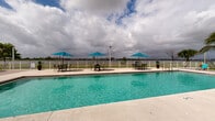 Located at 2201 N Australian Ave in West Palm Beach, Florida, this outdoor amenity area offers a resort-style experience defined by its generous swimming pool and scenic waterfront backdrop. The sparkling, crystal-clear pool is the centerpiece of the space, flanked by a wide, open concrete and wood-composite deck designed for relaxation and socializing. Vibrant aqua lounge chairs and patio umbrella sets are thoughtfully arranged throughout the deck, providing ample space for sunbathing and outdoor dining. A separate shallow wading area with step entry offers a more leisurely alternative alongside the main pool. Adjacent to the pool, a modern covered pavilion with a pergola-style roof provides a shaded lounge area, complemented by a glass-fronted clubhouse structure rendered in warm terracotta tones. Lush tropical palm trees and manicured greenery add a Florida resort feel throughout the outdoor space. A white perimeter fence frames the area while preserving sweeping, unobstructed views of the open waterway beyond, with the West Palm Beach skyline visible in the distance. A children's playground is also accessible nearby, making this a well-rounded outdoor community amenity suited for residents of all ages.