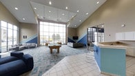 The Haven is a thoughtfully designed single-floor community space that combines comfort, style, and functionality. Upon entering, visitors are immediately drawn to the dramatic floor-to-ceiling windows that span an entire wall, bathing the interior in abundant natural light and offering open views of the surrounding exterior. The soaring vaulted ceilings enhance the sense of openness and airiness throughout the space.

The interior design features a cohesive color palette of blues, beiges, and whites, accented by bold geometric chevron wall detailing in blue and white that adds a modern, dynamic visual element. The flooring transitions from large-format tile near the entrance to a patterned, multi-toned carpet in the lounge areas, subtly defining different zones within the open layout.

A central pool table serves as a focal point for recreation and social interaction. Multiple seating arrangements with sofas and chairs are spread throughout, encouraging casual gatherings and relaxation. A counter-height bar area with modern stools provides an additional social hub. Recessed lighting and pendant fixtures complement the natural light, ensuring the space remains well-lit and welcoming at all hours. The Haven presents itself as an inviting and versatile gathering place.