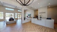 The Collins Clubhouse is a thoughtfully designed single-story community space that blends coastal charm with modern sophistication. The open-concept layout is anchored by striking herringbone-patterned hardwood flooring that flows throughout, complemented by white shiplap walls and soaring coffered ceilings with recessed lighting.

At the heart of the space, a welcoming reception desk greets visitors beneath a dramatic spherical iron chandelier. The lounge area near the entrance features an abundance of natural light streaming through floor-to-ceiling glass doors and clerestory windows, offering views of the outdoor pool area. A central seating zone with contemporary furnishings provides a versatile gathering spot.

A dedicated dining and refreshment area includes a dark wood communal table beneath an elegant crystal chandelier, accompanied by a built-in white cabinetry coffee bar with under-counter refrigeration. An adjacent lounge retreat features a floor-to-ceiling dark tile fireplace flanked by rustic wood floating shelves, plush seating, and a large multi-panel TV display mounted on a shiplap accent wall. Decorative organic wall art and carefully curated accessories throughout contribute to the warm, inviting coastal aesthetic of the space.