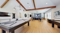 This single-level amenity lounge is designed for social gathering and recreation, offering a well-appointed and versatile space filled with natural light from large windows and glass doors. The architecture is defined by soaring vaulted ceilings accented with rich, exposed wood beams, lending the space a warm yet contemporary character throughout.

At the heart of the lounge sits a full-size pool table, positioned beneath a stylish multi-pendant light fixture. Adjacent to it, a shuffleboard table provides additional entertainment options, while a dedicated gaming station offers computer access. Comfortable seating areas are thoughtfully placed throughout, including casual lounge chairs and marble-top café tables ideal for small gatherings.

A generously sized island with a striking marble countertop and bar-height seating anchors the more social, collaborative zone of the room, illuminated by a sleek linear pendant light. Along the adjacent wall, a built-in wet bar with dark countertops and a mounted flat-screen TV creates a refined entertainment hub, complemented by a second wall-mounted TV nearby.

The space is finished with warm-toned luxury vinyl plank flooring that runs throughout, crisp white walls punctuated by slate-blue accent walls, and curated artwork. A round decorative mirror adds an elegant touch, while the overall design balances comfort and sophistication seamlessly.