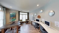 This thoughtfully designed single-floor space functions as a versatile workspace and lounge area, offering a welcoming environment suited for focused work and casual gatherings alike. The room features warm hardwood-style flooring that extends throughout, complemented by soft, light-toned walls that create a bright and open atmosphere. Recessed lighting in the smooth ceiling adds a clean, polished finish overhead.

A dedicated workstation area lines one wall, equipped with a long white desk and multiple computer monitors, providing ample room for individual or collaborative work sessions. Ergonomic rolling chairs complete the setup, ensuring comfort during extended use. Nearby, a rustic wooden table with bar-height seating offers an alternative workspace or informal meeting area.

Large windows dressed with elegant drape panels flood the interior with natural light, while a built-in bench seat beneath the windows creates a relaxed nook for reading or casual conversation. Decorative wall art, including a vibrant abstract painting and a curated arrangement of sculptural wall pieces, adds artistic character to the space. Lush greenery near the entrance introduces a refreshing natural element. The overall ambiance is modern, functional, and inviting — well-suited for a professional or community workspace setting.