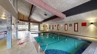 Located at 1920 NE Terre View Drive in Pullman, Washington, this single-floor indoor aquatic facility is purpose-built around a generously sized lap pool with clear, turquoise water. The pool is flanked by wide concrete pool decking that provides ample space for movement and safety. An adjacent hot tub/spa offers a relaxing complement to the main pool.

The facility features a vaulted ceiling supported by exposed dark wood beams, with large pink HVAC ducts running overhead — a functional architectural element that also adds industrial character. Walls are finished in a warm neutral tone and lined with a row of wood-framed windows that allow natural light to filter in. Accent lighting from wall-mounted sconces creates a welcoming ambiance throughout.

A standout feature is the pair of large glass-paneled garage-style doors on one end of the building, which open fully to connect the indoor pool area to an outdoor patio furnished with seating. This seamless indoor-outdoor transition allows for natural ventilation and views of the surrounding neighborhood. Entry doors and posted safety signage are clearly visible along the side walls. The overall design is utilitarian yet comfortable, making this space well-suited for both recreational swimming and structured aquatic activities.