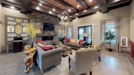 This single-floor clubhouse offers a thoughtfully designed communal space that blends rustic charm with modern comfort. The centerpiece of the main lounge is a dramatic floor-to-ceiling stacked stone fireplace, complemented by a wall-mounted television and framed by exposed wood beam ceilings with recessed lighting and an elegant chandelier. Plush gray sofas accented with bold red and yellow throw pillows create a warm, inviting seating arrangement, while wood-look tile flooring ties the space together with a natural, cohesive aesthetic.

The refreshment bar area is outfitted with white subway tile backsplash, dark wood cabinetry, quartz countertops, a stainless steel sink, and open shelving stocked with coffee and beverage amenities — all positioned beneath windows that allow natural light and outdoor views to filter in.

A dedicated business center area provides a functional workspace, featuring private room access, decorative shelving with curated accents, and casual seating at a striking red round café table. Framed artwork and sculptural wall décor enhance the character of this zone.

Throughout the clubhouse, the design maintains a cohesive rustic-modern personality, with carefully chosen furnishings, warm lighting, and layered textures that create an atmosphere that is both practical and welcoming for residents and guests alike.