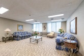 Located at 1477 Mississauga Valley Blvd in Mississauga, Ontario, this single-floor community space offers a well-appointed and versatile environment designed for both relaxation and social gathering. The space is organized into two distinct areas that complement each other seamlessly.

The lounge area is furnished with classic upholstered sofas and armchairs, accented by decorative artwork and floral arrangements. Patterned carpeting in neutral tones runs throughout, and large windows with vertical blinds allow ample natural light to fill the room, creating a warm and inviting ambiance. A mirrored wall feature adds a sense of depth to the space.

The dining area accommodates multiple round tables with cushioned metal chairs, providing flexible seating for gatherings or casual dining. Decorative framed artwork lines the walls, and lush indoor plants add a refreshing touch of greenery throughout.

A functional kitchenette services the space, equipped with warm wood cabinetry, a countertop with a built-in sink, a compact refrigerator, a microwave, and a toaster. Tile flooring in this area provides a practical and easy-to-maintain surface. Flush-mounted ceiling light panels provide consistent, even illumination throughout all areas of the space.