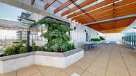 Located at 13639 George Junction in Surrey, BC, this rooftop amenity level offers a thoughtfully designed outdoor and indoor retreat high above the city skyline. The space is defined by its signature overhead wood pergola structure, whose warm timber beams provide partial shade across the generous tiled terraces while framing sweeping panoramic views of Surrey, the surrounding greenery, and distant mountain ranges through full-height glass railings.

The outdoor areas are carefully landscaped with raised concrete planter beds filled with mature trees, ornamental grasses, and shrubs, bringing a natural softness to the urban setting. Integrated bench seating lines the planters, and outdoor dining tables and chairs are positioned to take full advantage of the unobstructed views. The expansive terrace wraps around the building, offering multiple distinct zones for relaxation and gathering.

The indoor amenity area is finished to a high standard, featuring light wood laminate flooring, warm wood-panelled accent walls, and large windows that flood the space with natural light and maintain visual connection to the terrace greenery. A comfortable lounge area with a built-in media wall sits alongside a sleek kitchenette with grey cabinetry, a sink, and a casual dining nook. A glass-panelled door provides seamless access between the indoor and outdoor spaces, creating a cohesive and inviting amenity environment.
