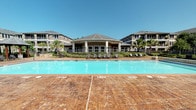Located at 4085 Hogan Dr in Tyler, Texas, this outdoor amenity space delivers a resort-inspired experience designed for relaxation and socializing. At the heart of the area is a large, sparkling swimming pool with a stamped concrete surround, flanked by rows of lounge chairs and outdoor dining sets, perfect for sunbathing or gathering with neighbors. A centrally positioned clubhouse pavilion with a distinctive hip roof and open-air columns serves as the visual anchor of the complex, providing shaded gathering space with direct pool access.

A beautifully constructed wooden gazebo with a stamped tile patio floor offers a comfortable retreat, furnished with wicker lounge seating and accent chairs cushioned in teal. This shaded seating area overlooks both the pool deck and the surrounding landscaped grounds, providing pleasant views of the manicured greenery, tropical palms, and ornamental shrubs that frame the community.

An outdoor kitchen station equipped with stainless steel grills and a stone-clad counter adds functional appeal, making the space ideal for outdoor entertaining. Wrought iron fencing encloses the grounds, contributing to both the aesthetic and the sense of community. The multi-story residential buildings surrounding the amenity area complete the setting, creating an inviting, well-appointed outdoor living environment.