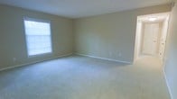 This single-floor space offers a straightforward and functional layout designed to maximize usability and comfort. The main room is generously sized, providing ample open floor space that lends itself well to a variety of furniture arrangements and living configurations. Soft, light-colored wall-to-wall carpeting covers the floor throughout, contributing to a warm and quiet atmosphere underfoot. The walls are finished in a neutral beige tone, creating a calm, versatile backdrop that pairs well with a wide range of décor styles. A window along one wall allows natural light to filter into the room, brightening the interior and adding a sense of openness. White baseboard trim lines the perimeter of the room, offering a clean and classic finishing detail. Electrical outlets are conveniently positioned along the walls to support practical everyday use. The space is presented in a clean, vacant condition, making it easy to envision and plan any desired furnishing or layout. The overall ambiance is understated and move-in ready, offering a blank canvas well-suited for residential use.