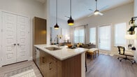 This single-floor residence showcases a thoughtfully designed open-concept layout that seamlessly connects the kitchen, dining, and living areas. The kitchen is a standout feature, centered around a large granite-topped island with seating for multiple guests and stylish pendant lighting overhead. Warm wood-tone cabinetry, stainless steel appliances — including a French door refrigerator and range — and wood-look flooring complete the contemporary aesthetic. Soaring ceilings amplify the sense of openness throughout the main living area, which is bathed in natural light from multiple large windows and a glass door leading to an outdoor area. A ceiling fan adds comfort to the bright and airy living space. The residence includes multiple bedrooms, each carpeted and well-lit by generous windows with blinds and curtains. The primary bathroom features a glass-enclosed walk-in shower and wood-tone cabinetry with granite countertops, while the secondary bathroom offers a full soaking tub and shower combination. Both bathrooms are finished with wood-look flooring and ample storage. The overall ambiance is modern and inviting, with a warm neutral palette, quality finishes, and a cohesive design sensibility throughout.
