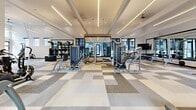 Located at 1801 E 121st Ave in Northglenn, Colorado, this fitness center offers a generously sized, single-floor exercise space designed to support a full range of workout routines. The open floor plan is finished with a distinctive checkered carpet tile pattern in neutral tones of gray and white, providing a clean, contemporary aesthetic throughout.

The space boasts soaring ceilings with linear LED lighting accents and ceiling fans, creating a bright and well-ventilated environment. Large windows along multiple walls and a full glass garage-style door flood the interior with natural light while offering views of the surrounding outdoor community areas.

Equipment is thoughtfully arranged to maximize usability. The cardio zone features treadmills, stationary bikes, a HIIT air bike, and rowing machines, while the strength training area is equipped with a variety of selectorized machines, free weights, a functional training rig with TRX suspension systems, and adjustable benches. Large mirrors are strategically placed to support proper form and visually expand the space.

The garage door opens directly to an outdoor courtyard, allowing for fresh air and a seamless indoor-outdoor connection. The modern, clean design combined with a comprehensive equipment selection makes this fitness center a well-appointed amenity for the community.