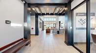This contemporary community clubhouse in North Aurora, Illinois, is designed to impress from the moment you enter. The grand lobby welcomes visitors with bold dark-stained exposed beam detailing overhead, striking dark structural columns, and large-format light tile flooring that extends throughout the space, creating a cohesive and polished aesthetic.

The open lounge area is a true centerpiece, boasting soaring double-height ceilings, a dramatic starburst chandelier, and floor-to-ceiling windows that flood the interior with natural light and offer views of the surrounding community. Comfortable seating arrangements — including a rich cognac leather sofa, upholstered chairs, and a stone-clad accent wall with a mounted flat-screen TV — establish an inviting and sophisticated atmosphere.

A built-in wet bar and cabinetry area with light wood finishes, open shelving, and a coffee station provide convenient amenities for residents and guests. The fully equipped demonstration kitchen features light wood cabinetry, stainless steel appliances, subway tile backsplash, white countertops, and industrial-style pendant lighting over a generous island with bar seating.

Dedicated dining areas offer both casual high-top seating and standard table arrangements, with large windows overlooking the outdoors. The space is thoughtfully designed to serve as a functional and stylish gathering hub for the community.