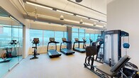 This fitness center offers a comprehensive exercise facility thoughtfully designed across a single open floor. The space is anchored by a generous cardio zone, featuring multiple treadmills, elliptical trainers, and stationary bikes—all positioned directly in front of expansive floor-to-ceiling windows that frame panoramic city views, creating an energizing workout environment flooded with natural light.

Beyond the cardio area, the facility includes a well-stocked strength training section equipped with a full dumbbell rack and various resistance machines. A glass-partitioned studio room provides a dedicated space for yoga, stretching, or group fitness activities, visible from the main floor to maintain an open and connected feel.

The facility's design is clean and modern, with a neutral color palette, light flooring, and a combination of recessed and linear LED lighting that ensures bright, even illumination throughout. Exposed ceiling elements add an industrial touch, while the white walls and glass partitions keep the space feeling airy and open.

Practical amenities include a dual water fountain station, a wall clock, and built-in cabinetry, all contributing to a functional and user-friendly environment. The overall ambiance is professional and inviting, making this fitness center a well-rounded destination for a full range of workout needs.