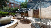 This property features a thoughtfully designed courtyard and interior common area that together create a welcoming and functional environment. The outdoor courtyard is paved with light-toned brick and furnished with multiple bistro-style metal tables and chairs arranged beneath large, slate-blue shade umbrellas. A curved fountain or planter border anchors one side of the patio, while mature potted palms and climbing vines along the perimeter wall add a lush, urban-garden feel. The courtyard is enclosed by a painted cinderblock wall and wooden gate, offering a semi-private retreat with views of the surrounding brick buildings and mature trees overhead.

The interior lobby and lounge area transitions smoothly from the entryway corridor, which features warm hardwood-style flooring, recessed lighting, and art pieces displayed along neutral gray walls. A console table and wall sconces provide functional elegance near the entrance. The lounge area is furnished with a cream-colored sofa, plaid accent chairs, and a wood coffee table arranged around a decorative teal niche displaying sculptural wall art. Bold illuminated marquee letters and colorful canvases add personality and visual interest throughout the space, contributing to an energetic yet comfortable ambiance.