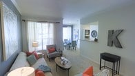 This single-floor residence features an open-concept design that connects the living and dining areas in a cohesive, functional layout. The living room is finished with plush wall-to-wall carpeting and neutral gray walls, creating a warm and inviting atmosphere. Large windows dressed with sheer white curtains allow generous natural light to fill the space, enhancing its airy feel. The living area comfortably accommodates a full sofa ensemble, accent chairs with coral throw pillows, and a central oval coffee table, providing both style and practicality.

The dining area sits adjacent to the living room and features a round table with upholstered chairs, ideal for everyday meals or entertaining. A pass-through window cutout in the wall connects the dining space to the kitchen, adding architectural interest while maintaining an open sightline. Decorative wall accents, including a circular medallion and framed artwork, add personality throughout.

Crown molding along the ceiling adds a refined architectural detail, elevating the overall finish of the space. The seamless flow between zones makes this a practical and well-appointed single-level home.