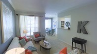 This single-floor residence features an open-concept design that connects the living and dining areas in a cohesive, functional layout. The living room is finished with plush wall-to-wall carpeting and neutral gray walls, creating a warm and inviting atmosphere. Large windows dressed with sheer white curtains allow generous natural light to fill the space, enhancing its airy feel. The living area comfortably accommodates a full sofa ensemble, accent chairs with coral throw pillows, and a central oval coffee table, providing both style and practicality.

The dining area sits adjacent to the living room and features a round table with upholstered chairs, ideal for everyday meals or entertaining. A pass-through window cutout in the wall connects the dining space to the kitchen, adding architectural interest while maintaining an open sightline. Decorative wall accents, including a circular medallion and framed artwork, add personality throughout.

Crown molding along the ceiling adds a refined architectural detail, elevating the overall finish of the space. The seamless flow between zones makes this a practical and well-appointed single-level home.