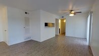 This single-floor residence offers a straightforward and functional layout designed for comfortable everyday living. The main living area is spacious and open, featuring light-toned hardwood-style flooring that extends throughout the space, contributing to a bright and cohesive aesthetic. Crisp white walls provide a clean, neutral backdrop, while a ceiling fan with integrated lighting adds both practicality and a subtle design element to the room. A defining architectural feature of the space is the built-in pass-through window connecting the living area to the kitchen, allowing for easy communication and an open feel between the two spaces. The kitchen, visible through the pass-through and the adjacent hallway, is warmly lit and equipped with essential appliances. Ventilation is well-addressed throughout, with visible ceiling vents and floor-level registers ensuring consistent airflow. A louvered door near the entrance hints at additional storage or utility space. The overall ambiance of this residence is tidy, understated, and move-in ready, making it well-suited for those seeking a clean, low-maintenance living environment with a practical and open interior flow.