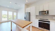 This well-appointed single-floor unit showcases a clean, contemporary design throughout, with crisp white walls and warm hardwood-style flooring that flows seamlessly from room to room. The open kitchen is a standout feature, outfitted with full-height white shaker cabinetry, a light gray tile backsplash, stainless steel appliances including a refrigerator with ice maker, range, microwave, and dishwasher, and a butcher block island that adds both function and character. Recessed lighting and a large glass door leading to the balcony ensure the kitchen and adjoining living area remain bright and inviting, with lush greenery visible beyond.

The bedrooms are well-proportioned and filled with natural light from large windows offering views of the surrounding neighborhood. One bedroom benefits from direct balcony access, extending the living space outdoors. The hallway connects the rooms efficiently, with additional doors providing closet and utility access.

The bathroom is finished to a high standard, featuring a wide double vanity with white cabinetry and quartz-style countertops, a large mirror with sconce lighting, and a frameless glass-enclosed walk-in shower with gray tile surround. The consistent use of light tones and quality finishes throughout lends the space a fresh, modern ambiance.