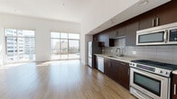 This contemporary single-floor residence showcases a thoughtfully designed open-plan layout that seamlessly blends the living and kitchen areas. Soaring ceilings and expansive floor-to-ceiling windows flood the generous living space with abundant natural light, framing impressive urban skyline views that bring the energy of the city indoors. The light-toned hardwood flooring extends throughout the main living area, enhancing the bright, airy atmosphere.

The kitchen is efficiently arranged along a single wall and features rich dark-walnut cabinetry contrasted by clean white countertops and a classic subway tile backsplash. A full suite of stainless steel appliances — including a gas range, over-the-range microwave, dishwasher, and refrigerator — provides a well-equipped cooking environment.

The bathroom is spacious and well-appointed, featuring large-format neutral floor tiles, a combination bathtub and glass-enclosed shower, a sleek dark-cabinetry vanity with a white countertop, and an adjoining water closet for added privacy. Built-in shelving within the bathroom offers practical storage.

Throughout the residence, a consistent modern design language is maintained through clean white walls, recessed lighting, and minimalist finishes, creating a refined and functional urban living environment.