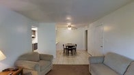 This single-floor space is characterized by an open-concept layout that seamlessly connects the living area, dining space, and kitchen. The living room features wall-to-wall carpeting, providing a comfortable foundation for seating, and transitions into the dining area, which is finished with square ceramic tile flooring. A ceiling fan with an integrated light fixture is centrally positioned overhead, serving both the dining and living zones while promoting air circulation throughout the shared space.

The dining area accommodates a modest table and chair set, situated between the living room and the kitchen. A partial wall acts as a natural divider between the dining space and the kitchen, which is visible in the background and features dark cabinetry and what appears to be standard kitchen appliances. A doorway on the far right side of the space leads to an additional room, suggesting further functional areas beyond the main open-plan zone. The walls throughout are painted in a neutral white, contributing to a clean, bright atmosphere. Overall, the layout is practical and straightforward, offering a cohesive flow between the main living areas.