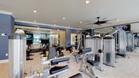 Located at 2330 Sawmill Place Blvd in Columbus, Ohio, this sophisticated fitness and wellness facility is thoughtfully designed to deliver a premium experience. The space is anchored by a expansive main gym area outfitted with a comprehensive selection of cardio equipment — including treadmills, ellipticals, and a StairMaster — alongside a robust array of strength-training machines and free weights. Deep navy accent walls, recessed lighting, ceiling fans, and mounted flat-screen TVs contribute to an energizing yet refined atmosphere throughout the workout area. A dedicated movement studio features herringbone hardwood flooring and a full wall-mounted equipment rack stocked with kettlebells, resistance bands, medicine balls, and TRX suspension systems, offering a versatile space for functional training. Floor-to-ceiling glass partitions connect the various zones while maintaining an open, airy feel. The common areas showcase dramatic black marble flooring with white veining, framed by wainscoting in rich navy blue and warm cream walls. A welcoming lounge nook provides a comfortable seating area with upholstered wingback chairs, decorative wall art, and soft ambient lighting. A serene massage room rounds out the wellness offerings, appointed with a treatment table, marble-topped cabinetry, and a softly curtained window for a calm, restorative environment. Elevator access and polished finishes throughout reinforce the facility's upscale character.