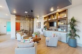 This thoughtfully designed single-floor space offers a seamless blend of modern aesthetics and functional living. The open-concept living and dining area serves as the heart of the space, anchored by light hardwood flooring that runs throughout. A striking vertical wood slat room divider acts as an architectural focal point, elegantly separating zones while maintaining visual continuity. The living area features a built-in shelving unit with warm wood cubbies, integrated cabinetry, and a media center, complemented by a bold dark-painted ceiling that adds depth and sophistication. A sculptural multi-arm pendant light fixture adds a designer touch overhead. Floor-to-ceiling windows dressed in sheer linen drapes flood the space with natural light and offer open outdoor views. The dining area is framed by vibrant abstract artwork and accommodates a generous table beneath a matching statement chandelier. The bedroom is carpeted and bright, featuring large windows with outdoor views and a vivid teal accent wall that energizes the room. An additional room with carpet and oversized windows provides flexibility for various uses. Throughout, the space strikes a balance between contemporary design and warm, inviting character.