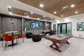 Located at 2000 James Jackson Pkwy in Atlanta, GA, this generously sized amenity lounge offers a dynamic, multi-zone recreational and social environment spread across a single open floor. The space is defined by its striking architectural details, including a slatted wood ceiling with recessed lighting that adds warmth and texture against crisp white walls and large-format light tile flooring.

The game area serves as a central focal point, anchored by a substantial pool table and complemented by a ping-pong table, arcade machines, and a shuffleboard table positioned against a bold geometric-patterned accent wall. A dedicated lounge zone features plush tufted velvet seating, modern curved sofas, and accent chairs arranged around coffee tables, creating an inviting space for relaxed socializing.

A media lounge area includes wall-mounted televisions backed by deep green glazed tile columns and a built-in wet bar with under-counter refrigeration, white countertops, and pendant lighting — ideal for gatherings. Additional seating vignettes with orange and rust-toned accent chairs provide casual dining and conversation spots throughout.

Curated contemporary artwork and thoughtful décor accents are displayed throughout the space, reinforcing a cohesive, upscale aesthetic. The overall ambiance is modern, energetic, and well-appointed, designed to accommodate a variety of social and recreational activities.
