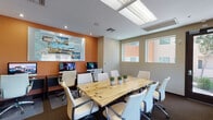 This single-floor professional space is thoughtfully designed to accommodate meetings, presentations, and collaborative work. The centerpiece of the room is a striking live-edge wood conference table surrounded by sleek white ergonomic chairs, creating a blend of natural warmth and contemporary style. Dark carpeting grounds the space, complementing the neutral beige walls and dark cabinetry positioned along the perimeter.

A dedicated workstation area along one wall features multiple desktop computers set against a bold terracotta accent wall, above which a large branded display board provides visual interest and context. This tech-equipped zone enhances the room's versatility for presentations and research.

Natural light flows generously through multiple windows fitted with roller shades, offering views of the surrounding exterior. A glass-paneled entry door adds to the brightness while providing direct outdoor access. Recessed and skylight-style overhead lighting ensures consistent illumination throughout.

Supporting amenities include a wall-mounted flat-screen television, a decorative table lamp atop a dark credenza, a printer station, and tasteful décor elements such as potted greenery and vase arrangements. A corridor connects to additional areas, suggesting the space is part of a larger professional facility.