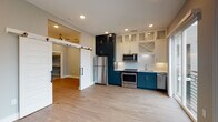 Located in Decatur, GA, this single-floor residence showcases a thoughtfully designed interior with a clean, contemporary aesthetic throughout. The open-concept kitchen is a standout feature, finished with a striking two-tone cabinet scheme — bold teal lower cabinets paired with crisp white upper cabinets, including glass-front and decorative X-detail doors that extend to the ceiling for ample storage. A full suite of stainless steel appliances, a white tile backsplash, and light countertops complete the functional and stylish culinary space. A sliding glass door adjacent to the kitchen fills the area with natural light and provides direct access to a private balcony with modern cable railings. A distinctive sliding barn door adds architectural character and connects the main living area to additional rooms. Light hardwood flooring runs throughout the living spaces, complementing the neutral gray walls and crisp white trim. The hallway features recessed lighting and panel-style doors that maintain the cohesive modern design. The bathroom is well-appointed with large-format stone-look tile lining the walls and floors, a soaking tub with chrome fixtures, and a curved shower rod. An additional flexible room with hardwood flooring offers versatile use as a bedroom or office.