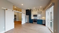 Located in Decatur, GA, this single-floor residence showcases a thoughtfully designed interior with a clean, contemporary aesthetic throughout. The open-concept kitchen is a standout feature, finished with a striking two-tone cabinet scheme — bold teal lower cabinets paired with crisp white upper cabinets, including glass-front and decorative X-detail doors that extend to the ceiling for ample storage. A full suite of stainless steel appliances, a white tile backsplash, and light countertops complete the functional and stylish culinary space. A sliding glass door adjacent to the kitchen fills the area with natural light and provides direct access to a private balcony with modern cable railings. A distinctive sliding barn door adds architectural character and connects the main living area to additional rooms. Light hardwood flooring runs throughout the living spaces, complementing the neutral gray walls and crisp white trim. The hallway features recessed lighting and panel-style doors that maintain the cohesive modern design. The bathroom is well-appointed with large-format stone-look tile lining the walls and floors, a soaking tub with chrome fixtures, and a curved shower rod. An additional flexible room with hardwood flooring offers versatile use as a bedroom or office.
