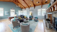 Located at 1140 Trinity Pine Ln in Raleigh, NC, this single-floor clubhouse is a beautifully appointed gathering space that blends rustic architectural charm with modern, stylish furnishings. The defining feature of the main lounge is its dramatic vaulted ceiling clad in warm natural wood planks, supported by exposed timber beams and trusses that draw the eye upward and create an airy, open atmosphere. Ceiling fans complement the soaring roofline, ensuring comfort throughout the space.

The main lounge area is anchored by a floor-to-ceiling stacked stone fireplace with a substantial wood mantel, serving as a striking focal point. Light hardwood flooring runs throughout, enhancing the bright, welcoming feel of the space. Soft blue-gray walls paired with teal and olive accent furnishings create a cohesive, contemporary coastal palette. Ample natural light pours in through numerous white-framed windows along multiple walls, offering views of the surrounding greenery.

Comfortable seating arrangements include a large sectional sofa, accent chairs, and a live-edge coffee table, all centered on a vibrant teal area rug. Decorative shelving units display curated accessories, adding personality and depth to the room. A separate billiards room provides dedicated entertainment space, complete with a classic pool table and additional natural light from corner windows. The overall ambiance is inviting, sophisticated, and thoughtfully designed for community gathering and recreation.