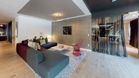 This single-floor amenity space is thoughtfully designed with multiple distinct zones that balance social activity and relaxed lounging. The central focal point is a dramatic double-sided fireplace clad in iridescent mosaic tile, flanked by dark built-in shelving displaying curated décor. Around it, hexagonal patterned floor tiles anchor a seating area furnished with sculptural teal wingback chairs accented with yellow cushions.

Light wood flooring extends throughout the remaining areas, leading to a bright, window-lined communal zone featuring a long bar-height table with industrial-style stools, bathed in natural light from floor-to-ceiling glazing with street views. Adjacent is a fully equipped kitchen island with a quartz countertop, integrated sink, and dark cabinetry, positioned near a lounge area with a large sectional sofa and wall-mounted television.

Additional zones include a moody lounge room with hexagonal wall detailing, layered seating in warm tones, and sheer curtained windows, as well as a game room with a pool table. A glass display case and art pieces punctuate the circulation corridors, lending a gallery-like quality throughout. Recessed lighting and carefully chosen furnishings create a cohesive, upscale atmosphere.