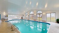 This expansive indoor aquatic facility is designed to provide a year-round swimming and relaxation experience. The centerpiece is a generously sized lap pool with crystal-clear turquoise water, accessible via multiple stainless steel entry rails and steps. A separate spa with its own dedicated enclosure and skylight structure is situated adjacent to the main pool, complete with posted capacity and rules signage, offering a distinct space for relaxation.

The architecture features soaring vaulted ceilings with recessed lighting that brighten the entire space. A striking row of arched windows along one wall, complemented by large sliding glass doors and additional rectangular windows, floods the area with natural light while providing open views of the surrounding outdoors. Teal accent trim around the windows and doors adds a distinctive decorative touch that ties the space together visually.

The pool deck is finished in neutral, light-toned tile and provides ample circulation space. A dedicated lounge and seating area features round tables with chairs, ideal for poolside relaxation. Safety equipment is conveniently stored on-site, and pool rules are clearly posted throughout. The overall ambiance is bright, clean, and welcoming, making this facility well-suited for both recreational swimming and leisure.