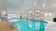 This expansive indoor aquatic facility is designed to provide a year-round swimming and relaxation experience. The centerpiece is a generously sized lap pool with crystal-clear turquoise water, accessible via multiple stainless steel entry rails and steps. A separate spa with its own dedicated enclosure and skylight structure is situated adjacent to the main pool, complete with posted capacity and rules signage, offering a distinct space for relaxation.

The architecture features soaring vaulted ceilings with recessed lighting that brighten the entire space. A striking row of arched windows along one wall, complemented by large sliding glass doors and additional rectangular windows, floods the area with natural light while providing open views of the surrounding outdoors. Teal accent trim around the windows and doors adds a distinctive decorative touch that ties the space together visually.

The pool deck is finished in neutral, light-toned tile and provides ample circulation space. A dedicated lounge and seating area features round tables with chairs, ideal for poolside relaxation. Safety equipment is conveniently stored on-site, and pool rules are clearly posted throughout. The overall ambiance is bright, clean, and welcoming, making this facility well-suited for both recreational swimming and leisure.