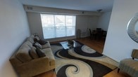 This single-floor residence offers an open and connected layout that integrates the living and dining areas into one cohesive space. The living area is anchored by warm hardwood flooring that extends throughout, providing a consistent and inviting foundation. A large picture window floods the room with natural light, fitted with horizontal blinds for adjustable privacy and light control. The living space comfortably accommodates a full-sized sofa and features a glass-top coffee table set atop a bold, swirling patterned area rug in tones of charcoal, tan, and cream — a striking focal point that adds character to the room. Transitioning seamlessly from the living area, the dining space is visible in the background and is furnished with a dark dining table and chairs, positioned near a doorway that suggests access to an adjacent room or hallway. The clean white walls and neutral color palette throughout create a bright, airy ambiance, while the open sightlines between the living and dining zones enhance the sense of spaciousness. The overall layout is practical and well-proportioned, making the space feel both functional and comfortable for everyday living.